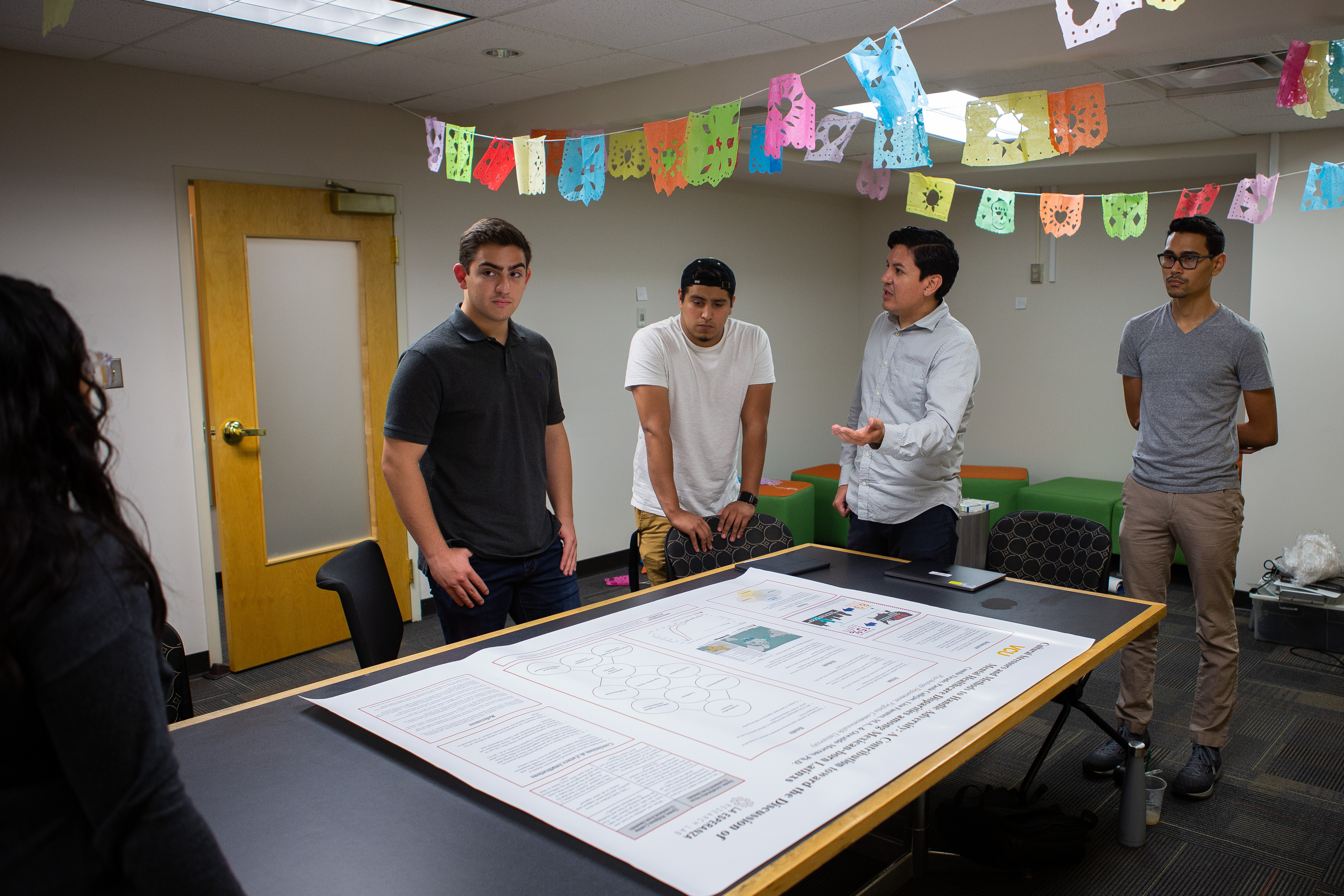 Students standing around a table with a large poster on it, discussing the contents.